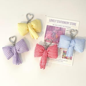 Puede incluir: Cuatro llaveros de lazo de vichy de colores con anillos en forma de corazón plateados. Los lazos son de color amarillo, morado, rojo y azul.