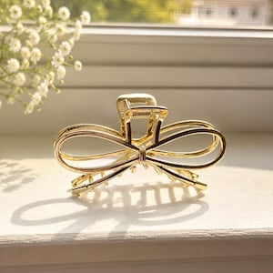 May include: A gold-toned metal hair claw clip featuring a symmetrical bow design with open loops. The accessory has a spring-loaded mechanism with interlocking teeth at the base to secure hair, finished with a polished, reflective metallic surface.