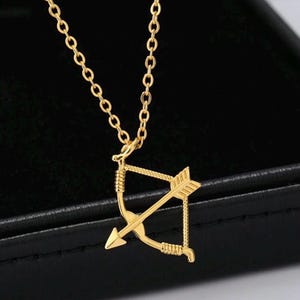 May include: A gold-colored necklace featuring a pendant shaped like a bow and arrow. The pendant is suspended from a delicate chain. The necklace is displayed against a dark background, highlighting the jewelry's details.