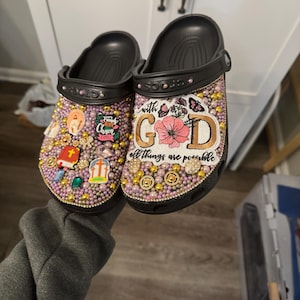 Könnte beinhalten: Ein Paar schwarze Crocs, verziert mit Strasssteinen, Perlen und Stoff-Patches. Die Patches zeigen religiöse Bilder, eine Blume und den Text "with God all things are possible".