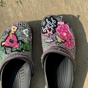 Könnte beinhalten: Ein Paar schwarze Clogs, verziert mit rosa, grünen und weißen Strasssteinen und verschiedenen Patches. Die Patches beinhalten einen rosa "Bad B*tch"-Patch, einen rosa Patch mit einem Comic-Gesicht und einen rosa Patch mit einem Dollarzeichen.