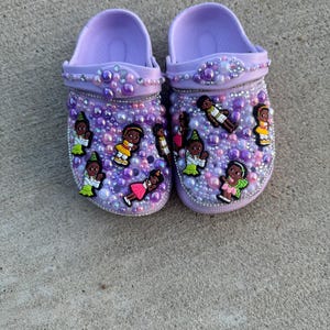 May include: A pair of lavender clogs decorated with colorful cartoon figures and iridescent beads.