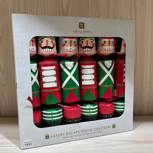 May include: A box of 12 Christmas crackers featuring a nutcracker design. The crackers are red, green, and gold with a white background. The box is white with the text "Talking Tables" and "Luxury Escape Room Crackers" printed on it. The text "Solve the puzzles to crack the code" is also printed on the box.