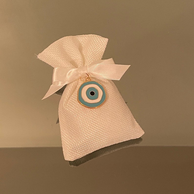K&ouml;nnte beinhalten: Eine kleine wei&szlig;e Stofftasche mit einem wei&szlig;en Satinband, das um die Oberseite gebunden ist. Ein goldener Anh&auml;nger mit einem blauen und wei&szlig;en b&ouml;sen Auge h&auml;ngt an der Tasche.