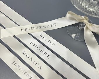 May include: Five off-white satin ribbons featuring black printed text. The ribbons are tied around the stem of a clear ribbed glass. The text on the ribbons reads BRIDESMAID, BRIDE, PHOEBE, MONICA, and JENNIFER in a clean, sans-serif font.