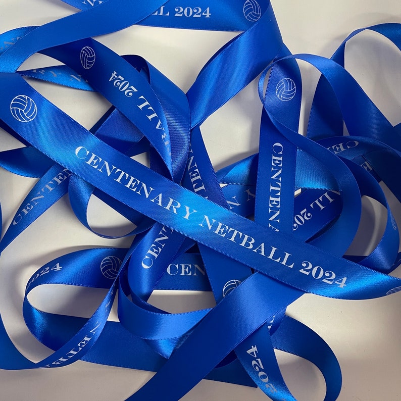 May include: Blue satin ribbon with the text "CENTENARY NETBALL 2024" and a netball graphic. The ribbon is tied in a knot and the ends are frayed.