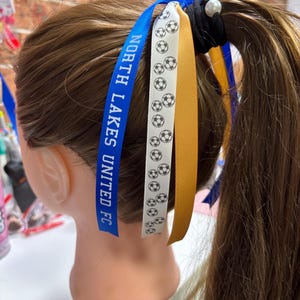 May include: A black hair tie with a white pearl accent featuring three attached ribbons. One ribbon is blue with the white text NORTH LAKES UNITED FC, one is white with a black football pattern, and one is a solid gold colour.