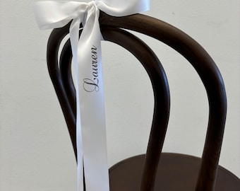May include: A white satin ribbon tied in a bow and attached to a dark wooden chair. The ribbon has the name "Lauren" printed in a script font. The chair has a curved back and a dark brown finish.