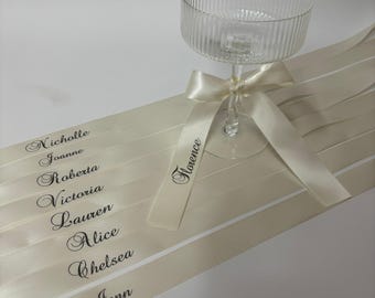 May include: A clear, ribbed glass coupe tied with a cream-colored satin ribbon. Several additional cream satin ribbons are laid out, each printed with a unique name in black script: Nicholle, Joanne, Roberta, Victoria, Lauren, Alice, Chelsea, Jenn, and Florence.