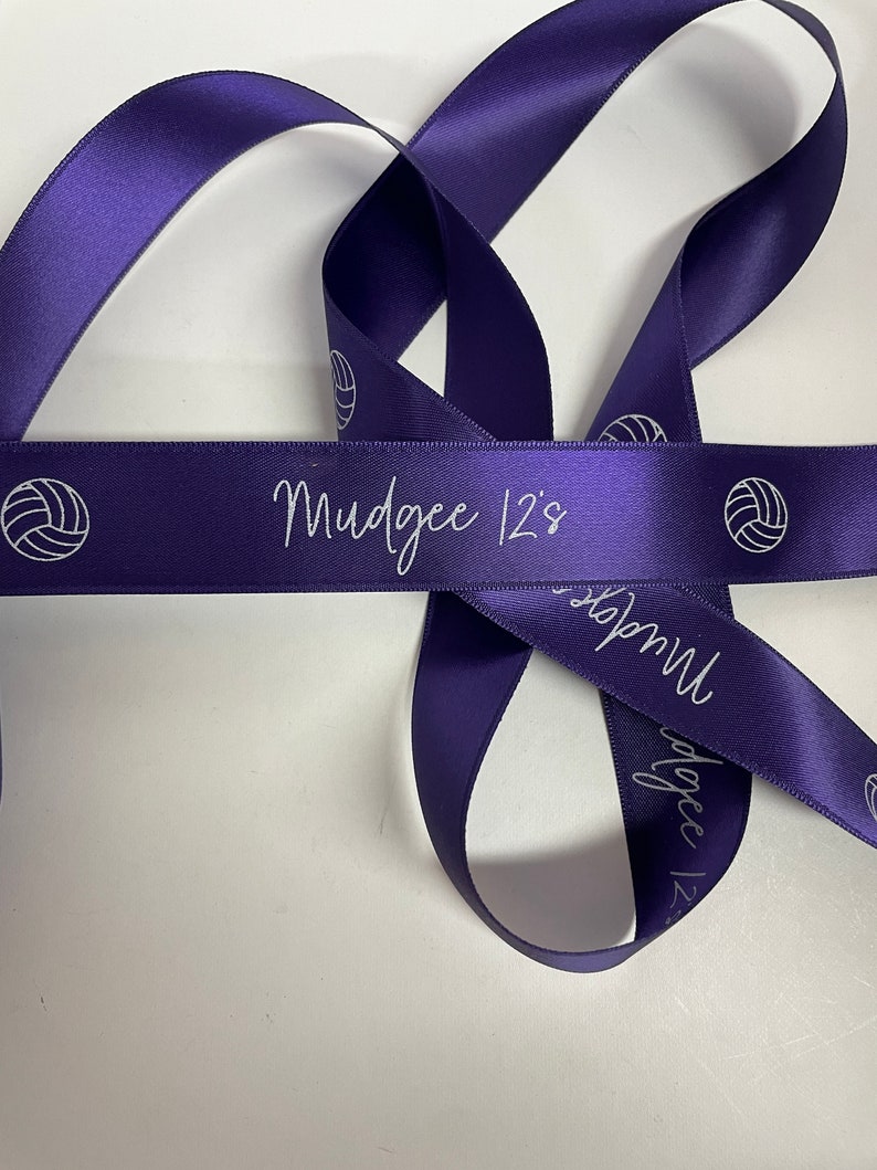 May include: Purple satin ribbon with white volleyball graphics and the text "Mudgee 12's" printed on it.