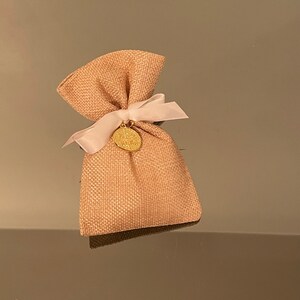 K&ouml;nnte beinhalten: Ein kleiner, beiger Jutesack mit einem rosa Band, das um ihn herum gebunden ist. Ein goldener Anh&auml;nger mit einem Design ist an dem Band befestigt.