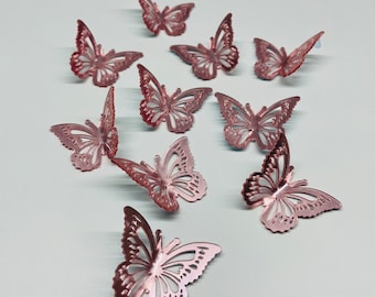 May include: A collection of rose gold butterfly decorations. The butterflies are made of metal and have intricate cut-out designs. They are arranged in a scattered pattern, suggesting a decorative theme.