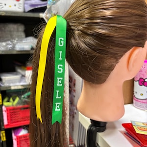 May include: A brown ponytail is held with a green ribbon printed with the name "GISELE" in white. A yellow ribbon is attached to the ponytail. The hair is styled on a mannequin head.