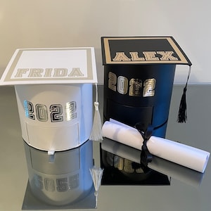 Peut inclure: Deux boîtes en forme de chapeau de graduation, une blanche et une noire, avec l'année "2022" et les noms "FRIDA" et "ALEX" en lettres argentées et dorées. Chaque boîte a un gland et un petit tiroir. Un diplôme roulé avec un ruban noir est devant la boîte noire.
