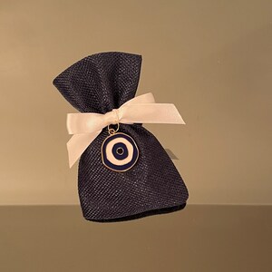 K&ouml;nnte beinhalten: Ein kleiner blauer und wei&szlig;er Augen-Anh&auml;nger h&auml;ngt an einem wei&szlig;en Band, das um eine dunkelblaue Jutesack gebunden ist.