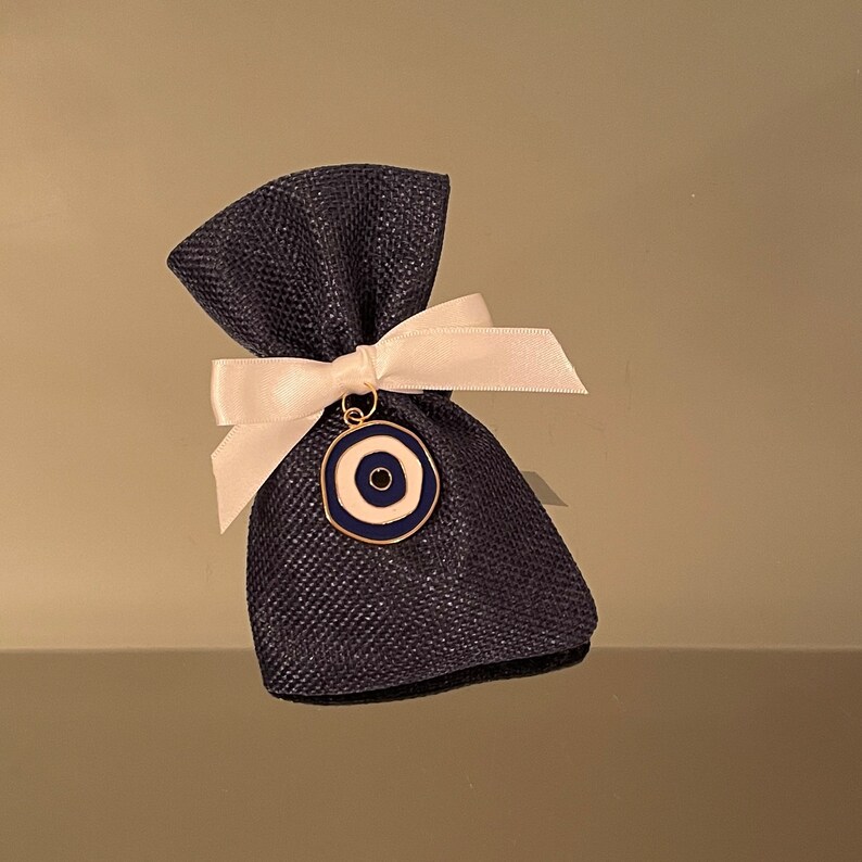 K&ouml;nnte beinhalten: Ein kleiner blauer und wei&szlig;er Augen-Anh&auml;nger h&auml;ngt an einem wei&szlig;en Band, das um eine dunkelblaue Jutesack gebunden ist.