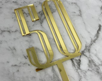 May include: Gold mirrored acrylic cake topper in the shape of the number 50. The number is designed with a modern, geometric style.