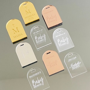 May include: Assortment of acrylic baby shower and baptism decorations. The decorations are in various colors, including gold, silver, and rose gold. The decorations feature text such as "Baby Shower", "The Arrival of", "Welcome", and "Baptism".