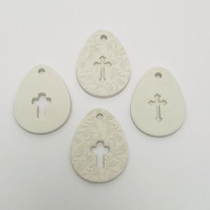 May include: Four white ceramic egg-shaped ornaments with a cross design. Two ornaments have a cross cut out, and two ornaments have a cross embossed with a floral pattern.