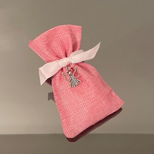K&ouml;nnte beinhalten: Eine kleine rosa Stofftasche mit einem wei&szlig;en Band, das um sie herum gebunden ist. Ein silberner Engelsanh&auml;nger h&auml;ngt an dem Band.