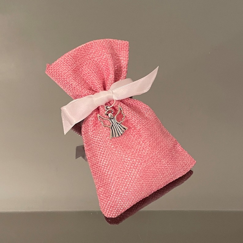 K&ouml;nnte beinhalten: Eine kleine rosa Stofftasche mit einem wei&szlig;en Band, das um sie herum gebunden ist. Ein silberner Engelsanh&auml;nger h&auml;ngt an dem Band.