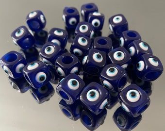 May include: A collection of blue and white evil eye beads. The beads are cube shaped and have a hole in the center for stringing. Each bead features a white evil eye with a blue pupil.