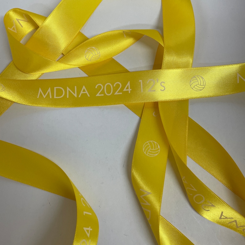 May include: Yellow satin ribbon with the text "MDNA 2024 12's" printed on it. The ribbon is folded and twisted, creating a decorative pattern.