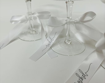 May include: Two clear glass coupes with a vertical ribbed texture on the bowls. Each glass features a white satin ribbon tied around the stem, with the name rachel printed in black cursive script on the trailing ends of the ribbon.