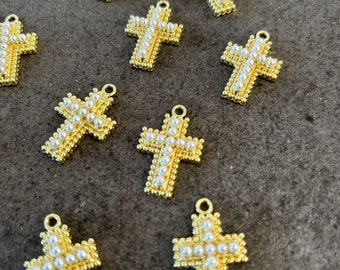 May include: A collection of ten gold-tone cross charms with white pearl accents. The charms are all the same size and shape, and they are arranged in a random pattern on a gray surface.
