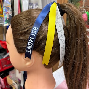 May include: A hair accessory featuring a black elastic band attached to three long satin ribbons. One ribbon is navy blue with the word BELMONT printed in white, one is solid yellow, and one is white with a repeating black basketball pattern.