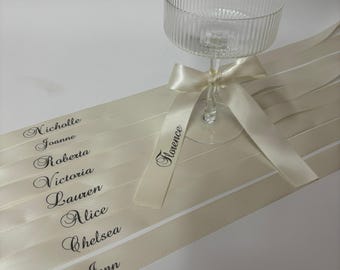 May include: A clear ribbed glass coupe with a stem, adorned with a cream-colored satin ribbon tied in a bow. The ribbon features the printed names Nicholle, Joanne, Roberta, Victoria, Lauren, Alice, Chelsea, Jenn, and Florence in black script.