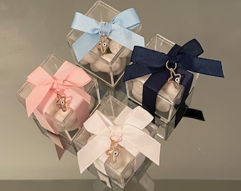 May include: Four clear, square boxes filled with white candies, each tied with a different colored ribbon: light blue, pink, navy, and white. Each ribbon is adorned with a gold-colored charm in the shape of a cross with an evil eye.