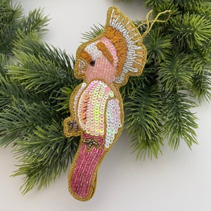 Peut inclure: Ornement de perroquet brodé de perles roses, dorées et de paillettes irisées. L'oiseau est bordé d'un fil d'or et orné de petites perles. Il est présenté sur un fond d'aiguilles de pin vertes.