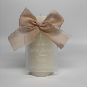 May include: A cylindrical white candle adorned with a beige frayed-edge ribbon bow. A translucent rectangular overlay features a white cross, the text GOD BLESS, Lola Isabella Jones, BAPTISED 2 JUNE 2023, God Parents, and EMILY BLUNT & JAMES JONES.