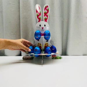 May include: A white bunny-shaped cardboard stand with blue and white foil-wrapped chocolate eggs. The stand has three tiers and is decorated with pink and green flowers.