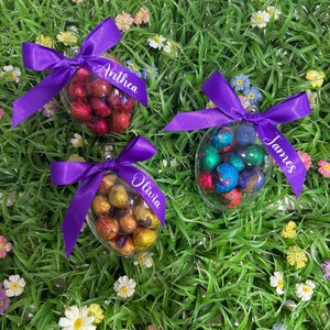 May include: Three clear, egg-shaped baubles filled with foil-wrapped chocolates, each tied with a purple ribbon. The ribbons are labelled with the names "Anthea", "Olivia", and "James". The baubles are set on a bed of green grass with small flowers.