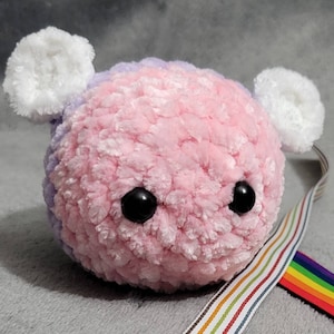 May include: A pink and purple crocheted plush toy with large white ears and black eyes. The toy is made of soft, fuzzy yarn and has a sweet, friendly expression.