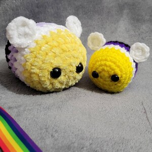 May include: Two crocheted bee plushies, one larger and one smaller. The larger bee is yellow and white with purple accents, while the smaller bee is yellow with white and purple stripes. Both bees have black button eyes.
