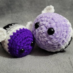 May include: Two crocheted plush toys shaped like bees with black eyes and white wings. One bee is purple and white, and the other is lavender and white.