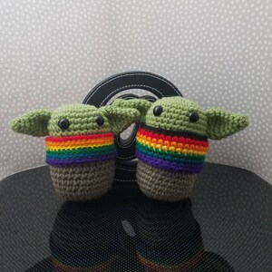 May include: Two crocheted green figures with rainbow colored bands around their necks. The figures are sitting on a black surface.