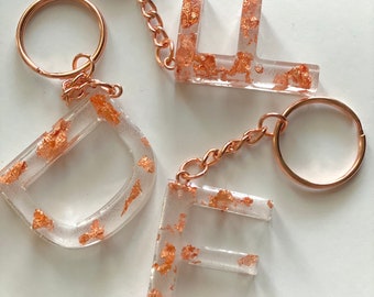 Op de afbeelding: Drie transparante sleutelhangers van hars met gouden vlokken. Elke sleutelhanger heeft een andere letter: D, E en E. De sleutelhangers zijn bevestigd aan gouden sleutelringen.
