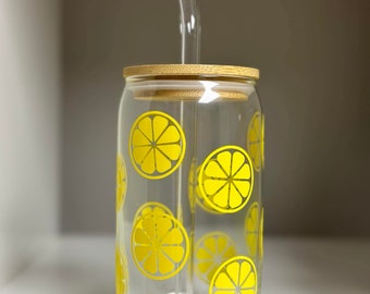 May include: A clear glass reusable water bottle with a bamboo lid and a yellow lemon design. The bottle has a glass straw.