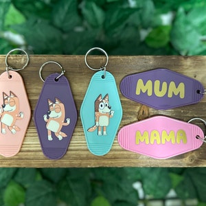 May include: Five keychains featuring a cartoon dog. Two keychains are pink and say "MAMA" and "MUM" in gold lettering. The other three keychains feature the cartoon dog in different poses on a purple, blue, and pink background.