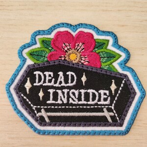May include: Embroidered patch featuring a black coffin shape with the words "DEAD INSIDE" in white. A pink flower with green leaves sits atop the coffin. The patch has a blue and white border, suitable for jackets, bags, or other fabric items.