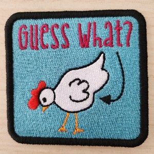 May include: Embroidered patch with a blue background and black border. The patch features a white chicken with a red comb and a black tail feather. The text "GUESS WHAT?" is embroidered in pink above the chicken.
