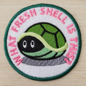 May include: A white embroidered patch with a green turtle inside a pink shell. The text "What Fresh Shell Is This?" is written around the turtle in pink.