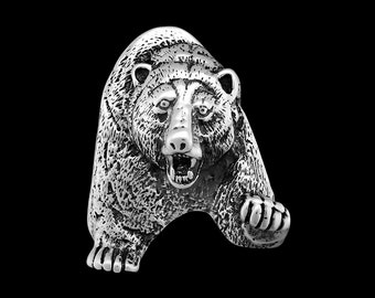 May include: A silver bear ring with a detailed, textured design. The bear is depicted with an open mouth and sharp teeth, giving it a fierce and menacing appearance.