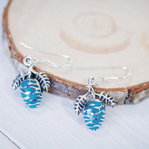 May include: A pair of silver dangle earrings with blue and silver pine cone charms. The pine cones have a textured surface and are accented with small silver leaves.