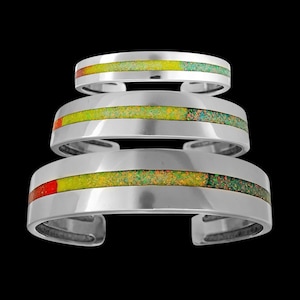 Könnte beinhalten: Drei silberne Armbänder mit grünem, gelbem und rotem Einlegearbeiten. Die Armbänder sind übereinander gestapelt.