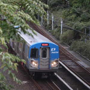 May include: A blue and silver PATH train with the word "NEWARK" displayed on the front, travelling on railway tracks.